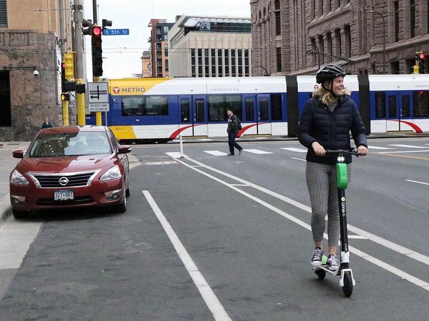 Outside City Hall, A light rail train passes a pedestrian and person riding a scooter