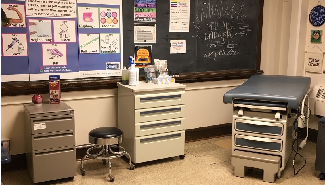 Room with medical exam table, stool and cabinets on wheels, blackboard and posters