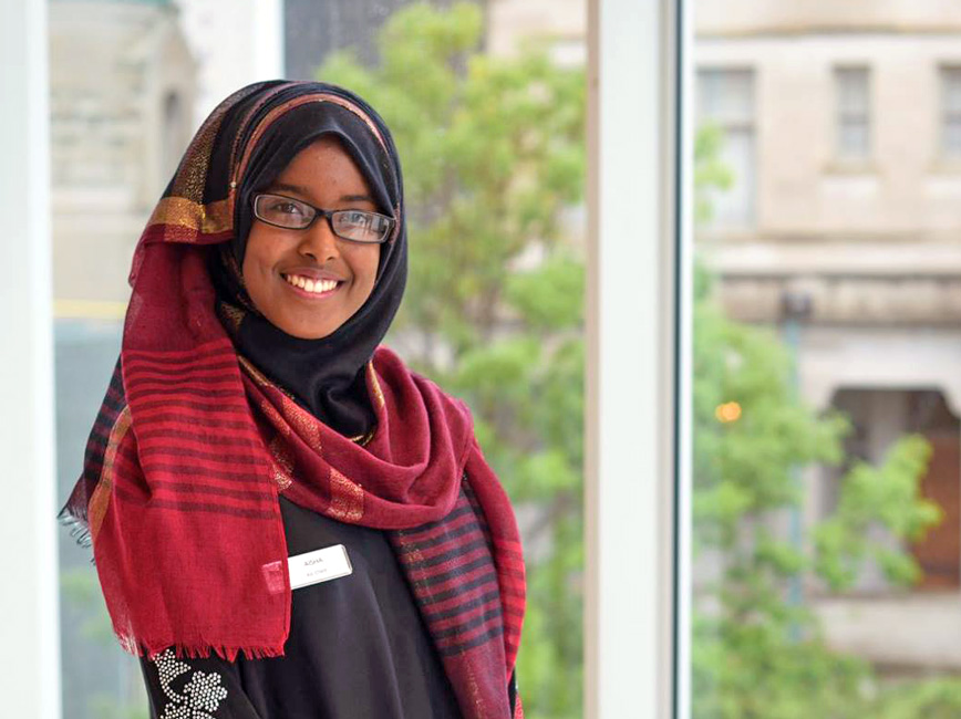 Smiling young person wearing glasses and colorful garments standing by window.