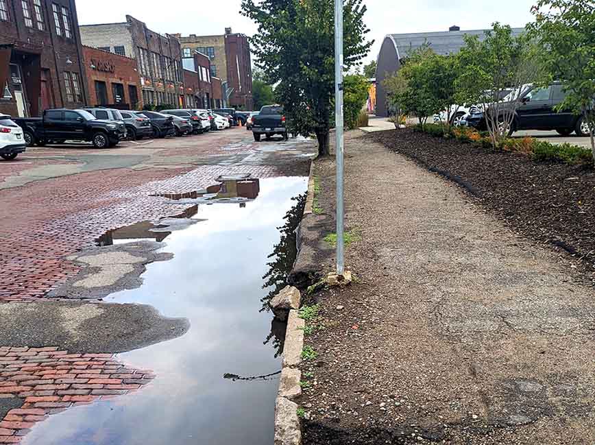 The edge of a brick paved street showing pooled water, a concrete curb broken into many pieces and a deteriorated asphalt sidewalk. The edge of a brick paved street showing pooled water, a concrete curb broken into many pieces and a deteriorated asphalt sidewalk.